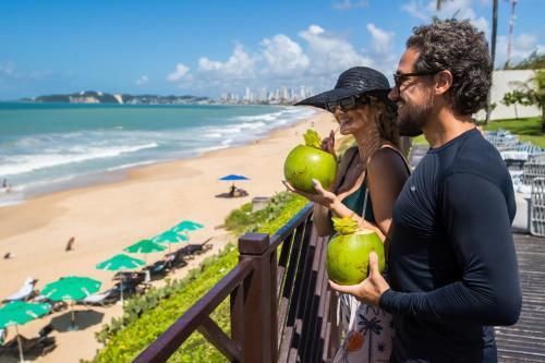 Duas pessoas em uma varanda na praia segurando cocos, com vista para uma costa ensolarada e água turquesa.