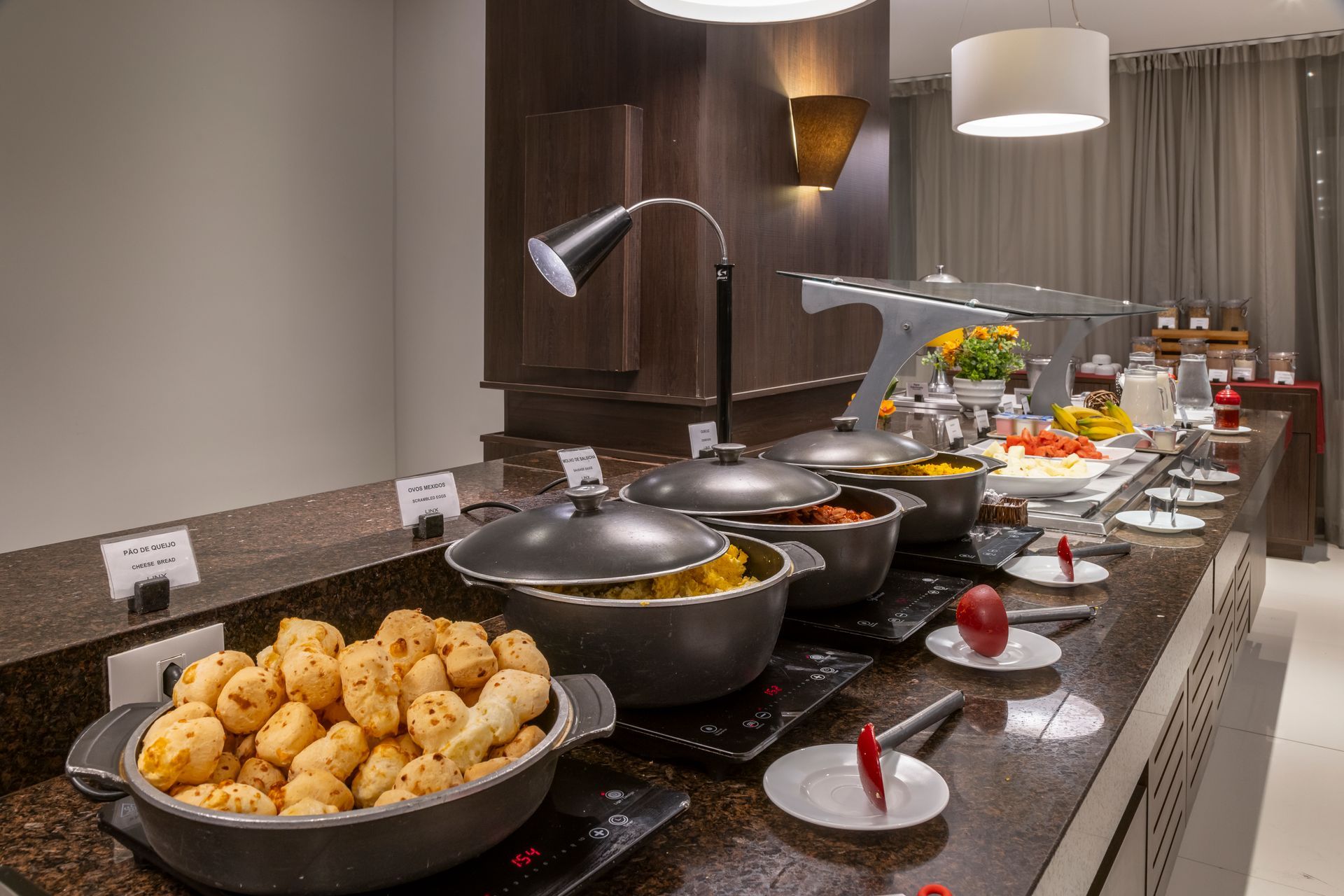 Buffet de hotel com tigelas de comida e frutas em um balcão de granito sob luzes aconchegantes.