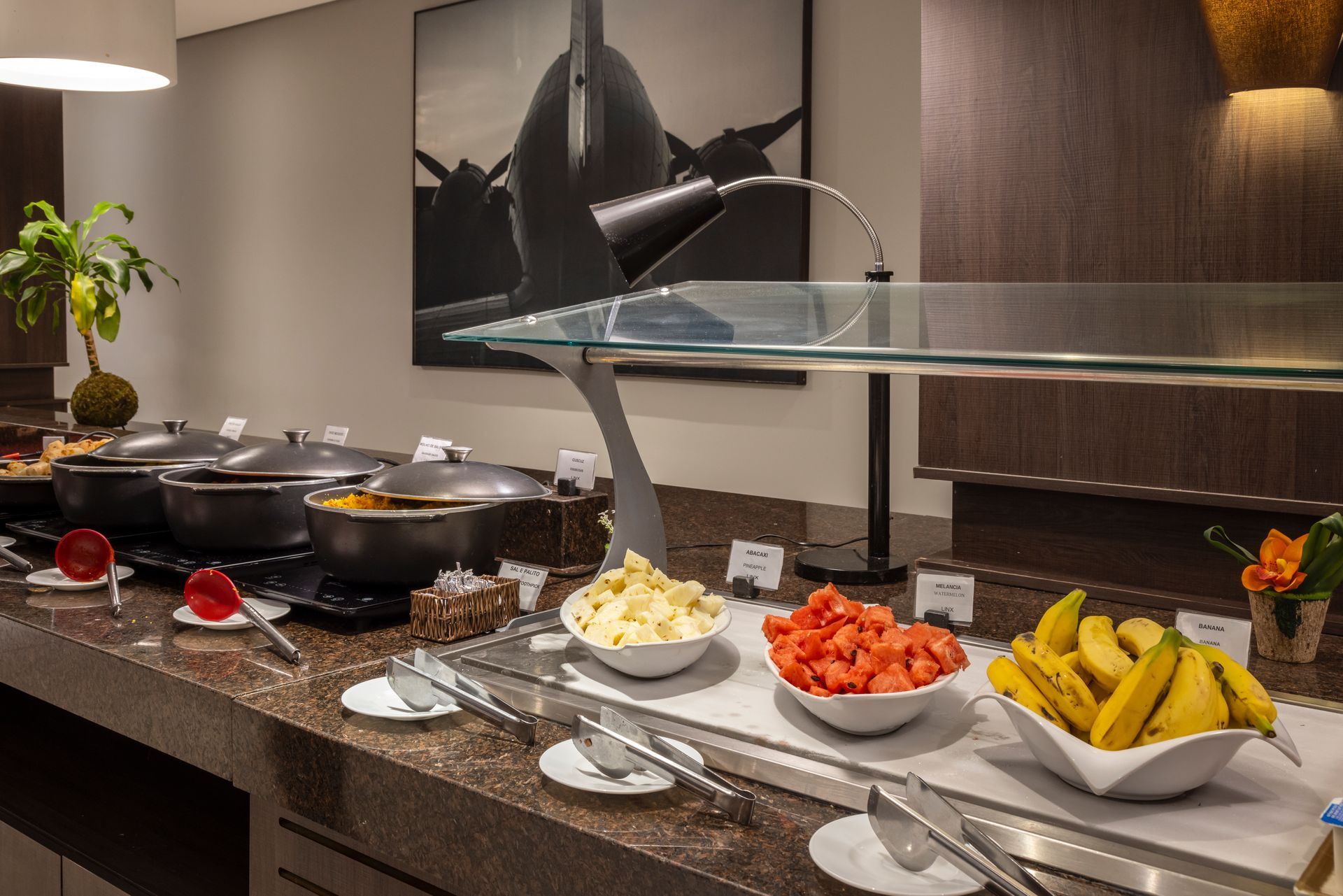 Buffet de hotel com bandejas de frutas, queijos e sobremesas sob uma vitrine de vidro.