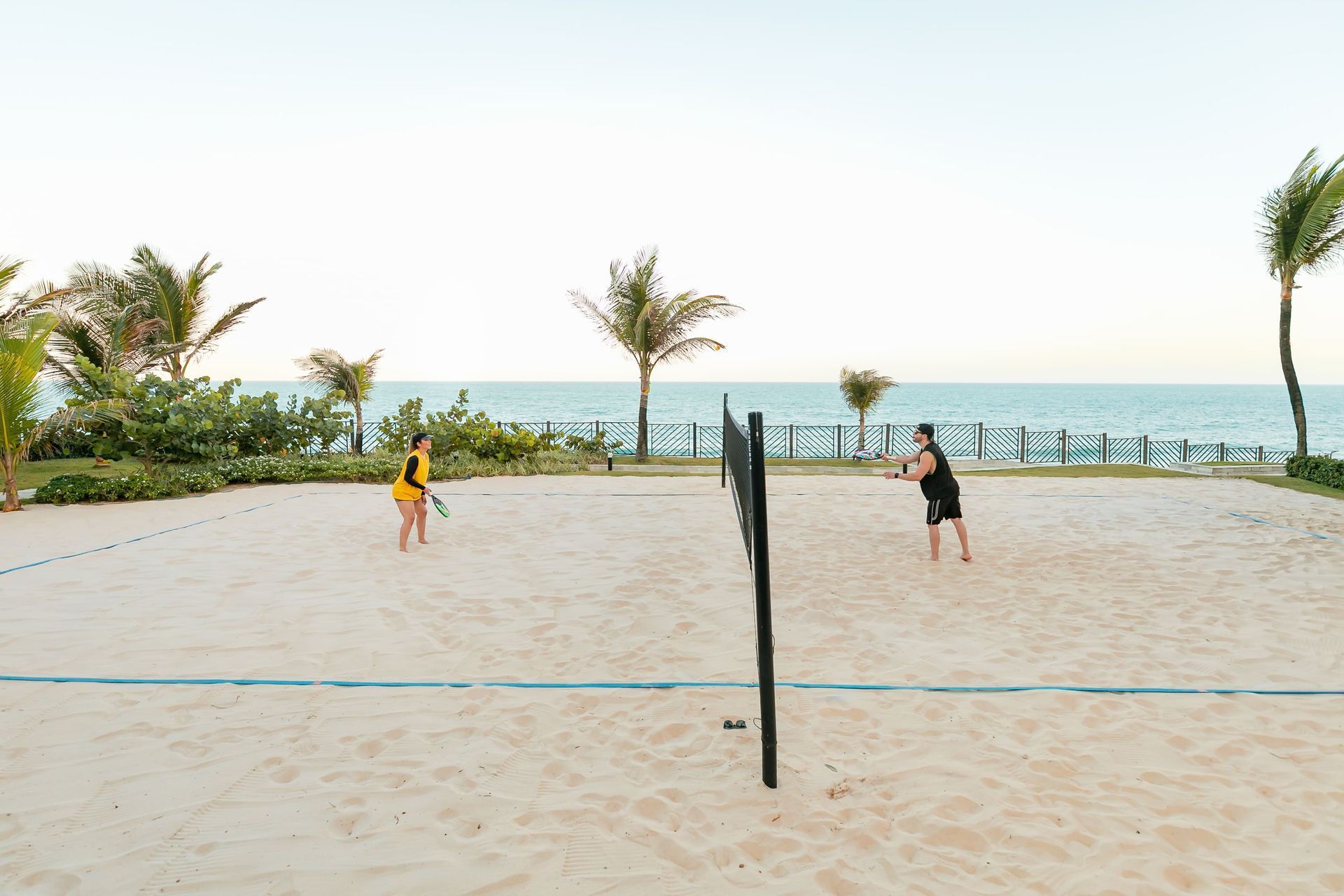 Duas pessoas jogando vôlei de praia em uma quadra de areia à beira-mar, com palmeiras e céu claro.
