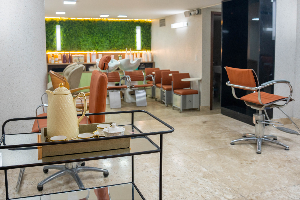 Salão de beleza moderno com cadeiras laranja, mesas de vidro e balcão com iluminação verde.
