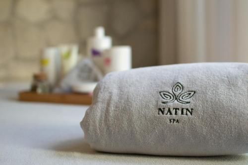 Toalha de hotel branca dobrada com o logotipo “NATIN SPA” sobre uma cama; artigos de higiene pessoal desfocados ao fundo.