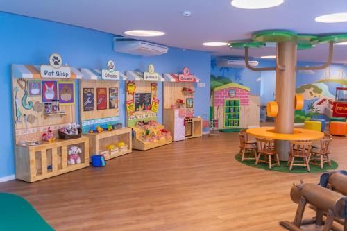 Sala de jogos pré-escolar colorida com lojas de brinquedos, mesa redonda, cadeiras e piso de madeira.