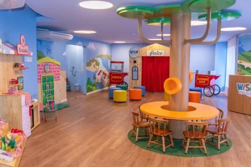 Sala de jogos infantil ampla e colorida, com mesas, cadeiras, prateleiras e um palco com cortina vermelha.