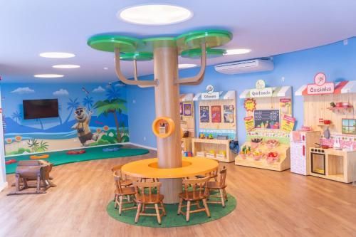 Sala de jogos com mesa central em formato de árvore, paredes coloridas, cozinha de brinquedo e pequena área de estar.