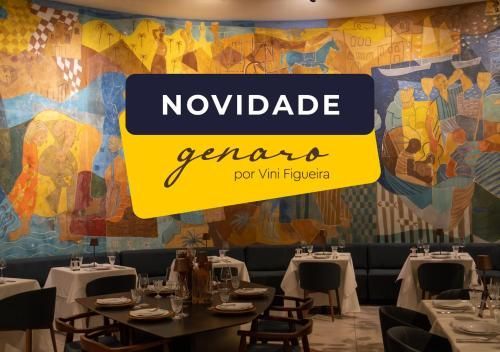 Faixa com a palavra “NOVIDADE” sobre um mural de restaurante em tons de amarelo e azul; mesas dispostas em primeiro plano.