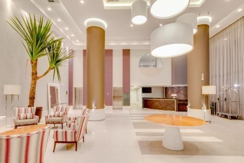 Sala de estar moderna e luminosa com colunas brancas e douradas, mesas redondas e uma palmeira em vaso, em um interior minimalista.