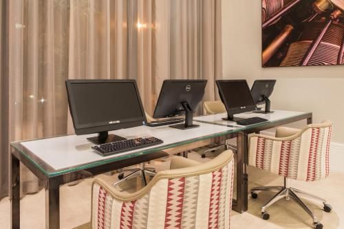 Espaço de trabalho moderno com três computadores de mesa em uma bancada comprida e banquetas listradas em uma sala com cortinas.