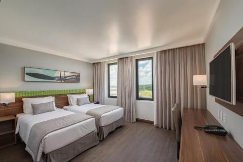 Quarto de hotel moderno com duas camas, TV de parede, janela e cortinas bege.