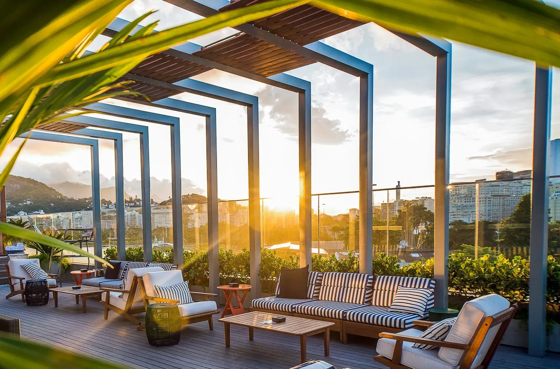 Um terraço ensolarado com cadeiras listradas, mesas e pérgola emoldura a vista da cidade ao pôr do sol.