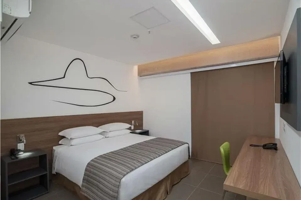 Quarto de hotel moderno com cama de casal, escrivaninha de madeira, cadeira verde e arte abstrata na parede.