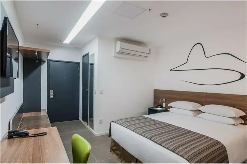 Quarto de hotel moderno com cama, escrivaninha, cadeira verde, obras de arte na parede e porta do banheiro.