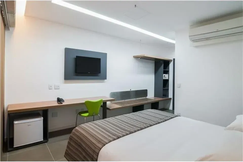Quarto de hotel moderno com cama, escrivaninha, TV de parede, cadeira verde e ar-condicionado.