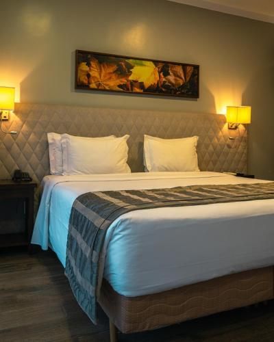 Quarto de hotel com cama arrumada, travesseiros brancos, luminárias de parede e obras de arte emolduradas acima da cabeceira.