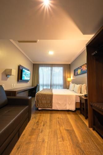 Quarto de hotel pequeno e moderno com cama, sofá, escrivaninha, TV e janela grande com cortinas.