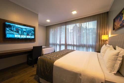 Quarto de hotel moderno com cama, TV de parede, escrivaninha e janela grande com cortinas translúcidas.