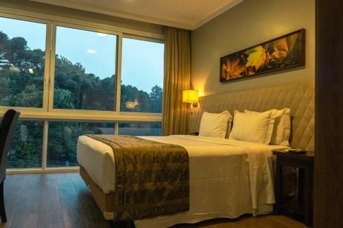 Quarto de hotel aconchegante com cama arrumada, abajur com luz quente e janela ampla com vista para as árvores ao entardecer.