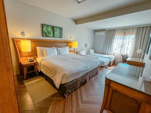 Quarto de hotel com duas camas, luminárias aconchegantes, móveis de madeira e janela com cortinas.