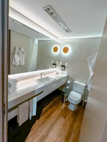 Banheiro moderno com bancada longa branca, espelho, piso de madeira e vaso sanitário sob luminárias de parede.