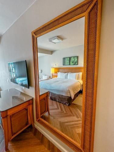 Espelho de parede com moldura de madeira refletindo um quarto com cama e piso de madeira em espinha de peixe.