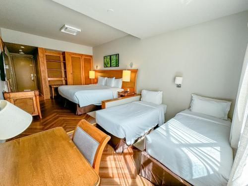 Quarto de hotel luminoso com três camas de solteiro, móveis de madeira e luz solar entrando pela janela.