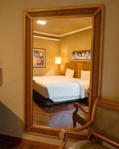 Quarto visto em um espelho de parede, com uma cama arrumada, um abajur, um quadro e uma cadeira em primeiro plano.