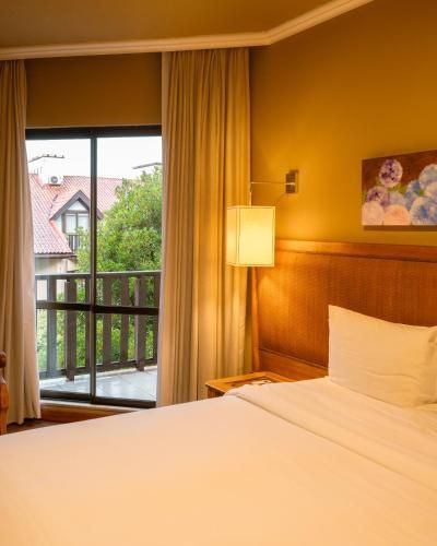 Quarto de hotel com cama arrumada, abajur, porta para varanda e vista para uma área verde externa.