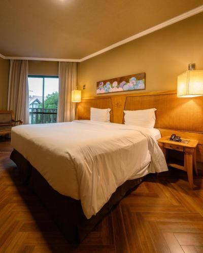 Quarto de hotel aconchegante com uma cama grande, cabeceira de madeira, abajures e janela com cortinas.