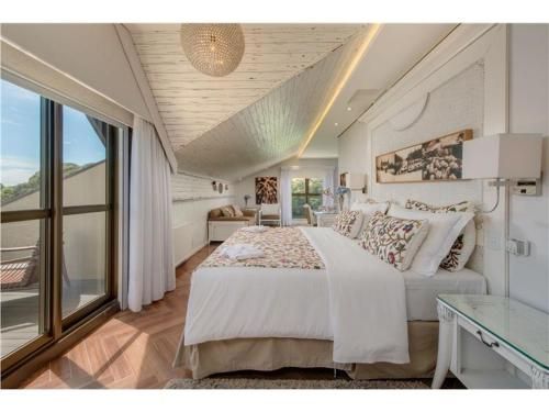 Quarto moderno e luminoso com janelas amplas, roupa de cama branca e teto inclinado de madeira com vista para uma varanda.
