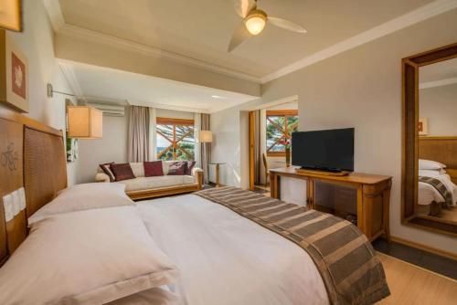 Quarto de hotel luminoso com duas camas, TV, móveis de madeira e grandes janelas com vista para a vegetação.