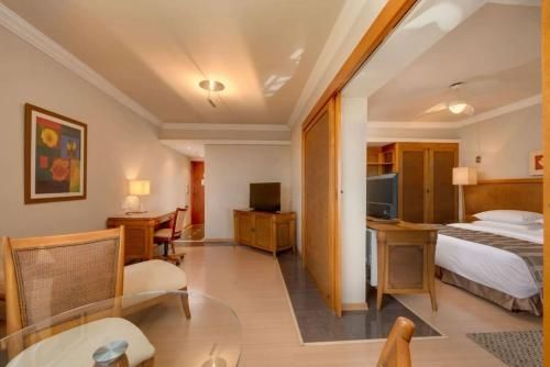 Suíte de hotel com decoração em tons de bege, área de estar, escrivaninha e quarto visível através de portas de correr abertas.