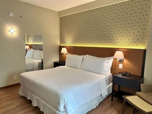 Quarto de hotel com uma grande cama branca, parede com estampa decorativa, abajures e uma cadeira junto à janela.