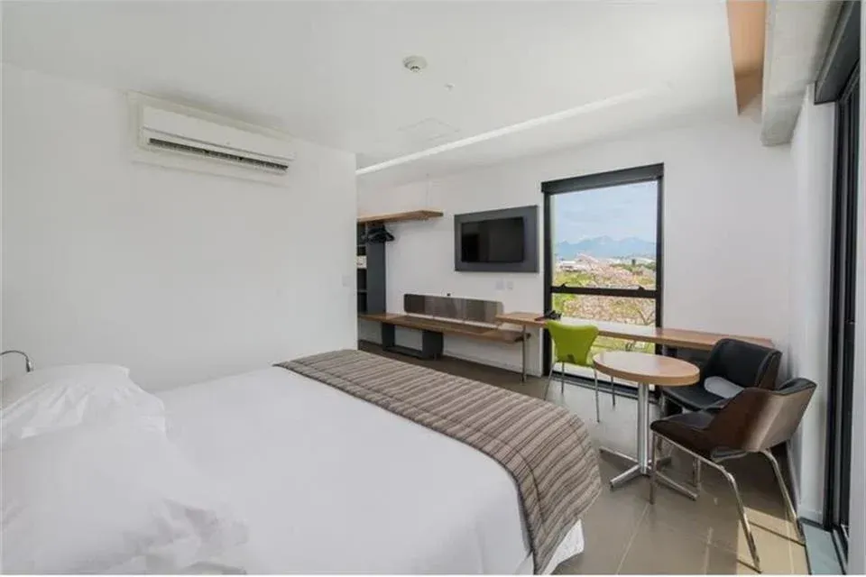 Quarto moderno com cama, TV, escrivaninha e janela ampla com vista para as montanhas.