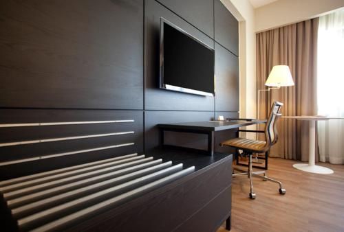 Quarto de hotel moderno com escrivaninha de madeira escura, TV de parede, cadeira de escritório e luminária de chão junto à janela.