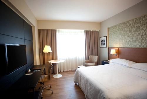 Quarto de hotel moderno com cama, TV, luminária, cadeira e cortinas claras na janela.