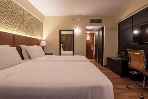 Quarto de hotel moderno com duas camas brancas, escrivaninha, cadeira, espelho e móveis de madeira escura.