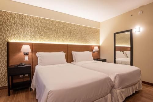 Quarto de hotel com duas camas de solteiro, iluminação aconchegante, abajures e espelho de parede.