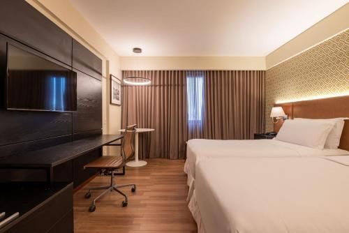 Quarto de hotel moderno com cama king-size, escrivaninha, TV e cortinas do chão ao teto.