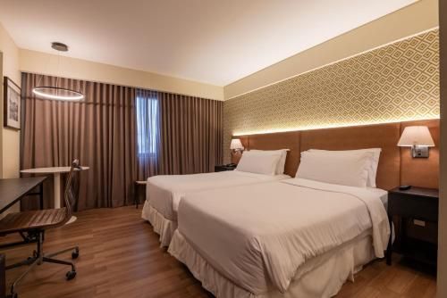 Quarto de hotel moderno com duas camas brancas, iluminação aconchegante, escrivaninha e janela com cortina.