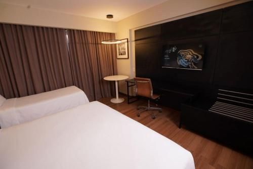 Quarto de hotel moderno com cama grande, cadeira de escritório, TV, cortinas e iluminação aconchegante.