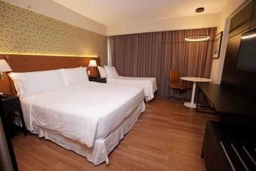 Quarto de hotel moderno com cama grande, piso de madeira, escrivaninha e TV de parede.