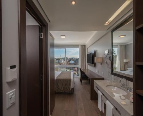 Quarto de hotel moderno com cama, escrivaninha, lavatório no banheiro e ampla janela com vista para a cidade.