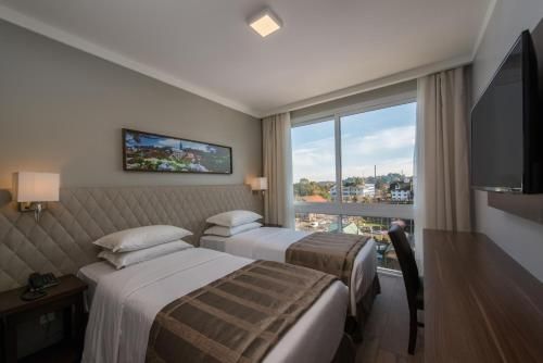 Quarto de hotel com duas camas bem arrumadas, uma escrivaninha, TV de parede e uma grande janela com vista para a cidade.