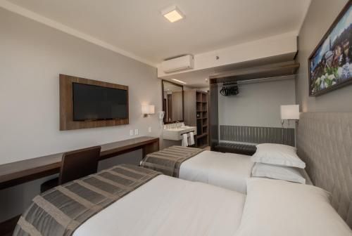 Quarto de hotel moderno com duas camas, TV de parede, escrivaninha e janela ampla com vista para a cidade.