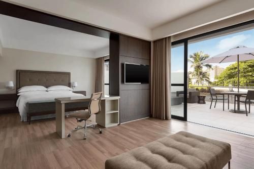 Quarto de hotel moderno com cama king-size, escrivaninha, TV e portas de correr que dão para um pátio ensolarado.