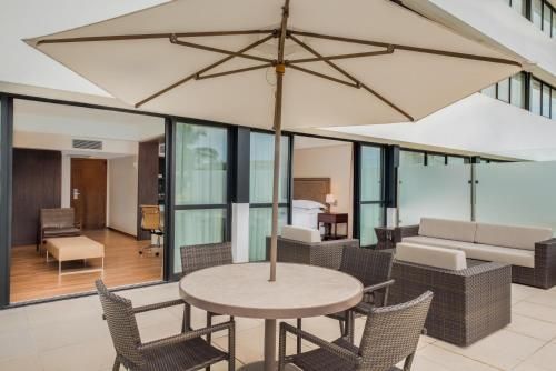 Pátio com mesa redonda, cadeiras, sofá e grandes guarda-sóis bege em frente a um apartamento moderno.