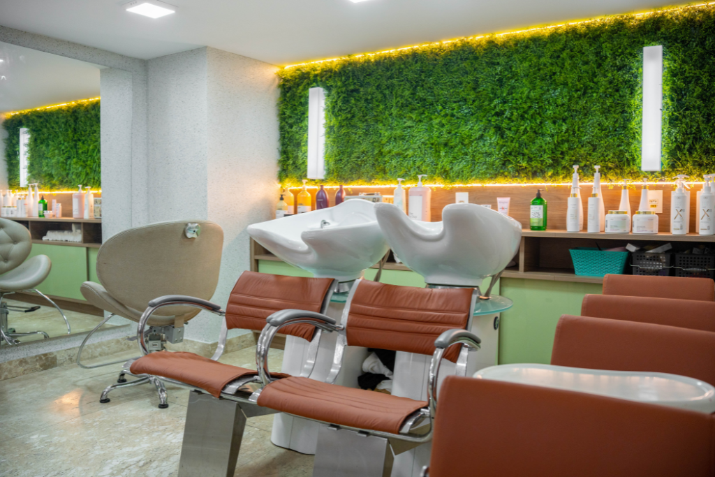 Área de espera moderna do salão de beleza com cadeiras reclináveis, espelhos e uma parede verde com plantas atrás das estações de trabalho.