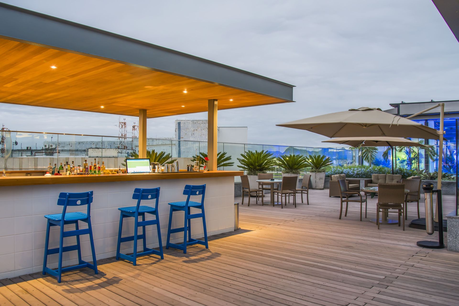 Bar ao ar livre na cobertura com banquetas azuis, balcão de madeira e sofás para relaxar ao pôr do sol.