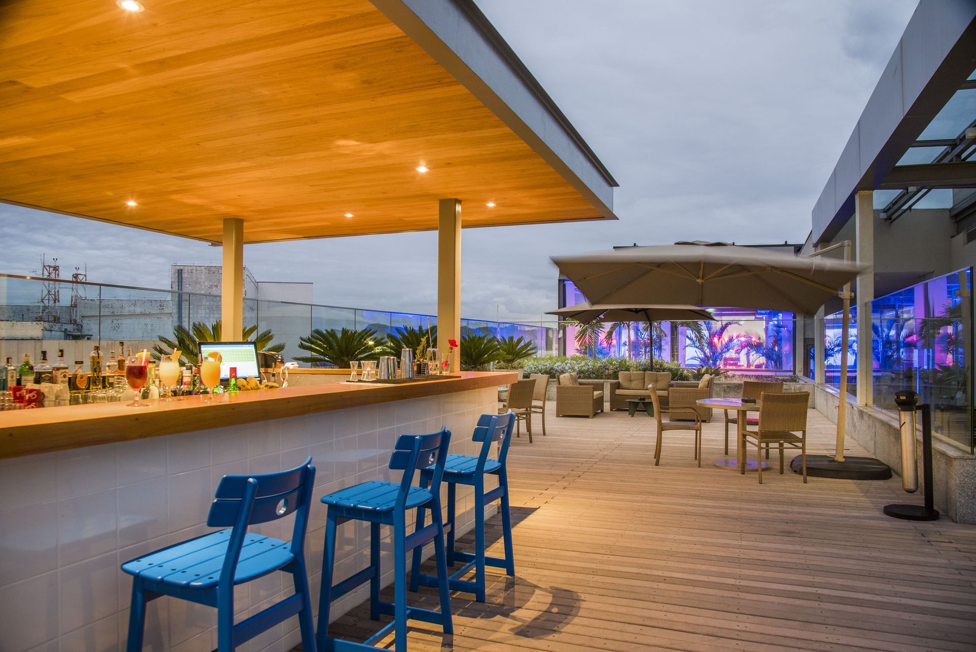 Terraço com bar na cobertura, cadeiras azuis, cobertura de madeira e luzes da cidade ao entardecer.
