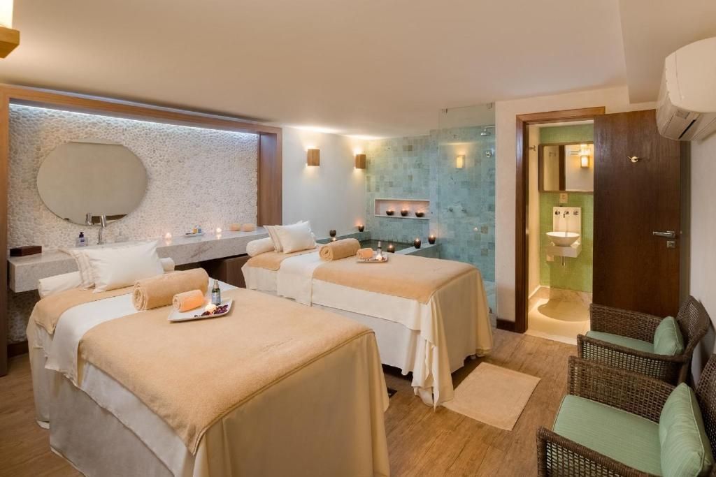 Sala de tratamento de spa com duas mesas de massagem, iluminação aconchegante e área de ducha revestida com azulejos.